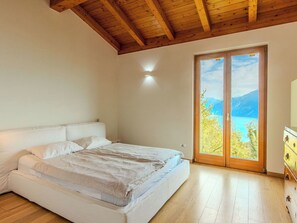 A bedroom with a bed, wooden flooring, a ceiling with exposed beams, a wall-mounted light, and a sliding glass door leading to a scenic view.