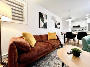 A modern living room with a brown sofa, two yellow cushions, a round coffee table, and a remote control on it. - Stylish 3BR Condo for Monthly Stays | 2 Baths, Laundry & AC (Montréal)