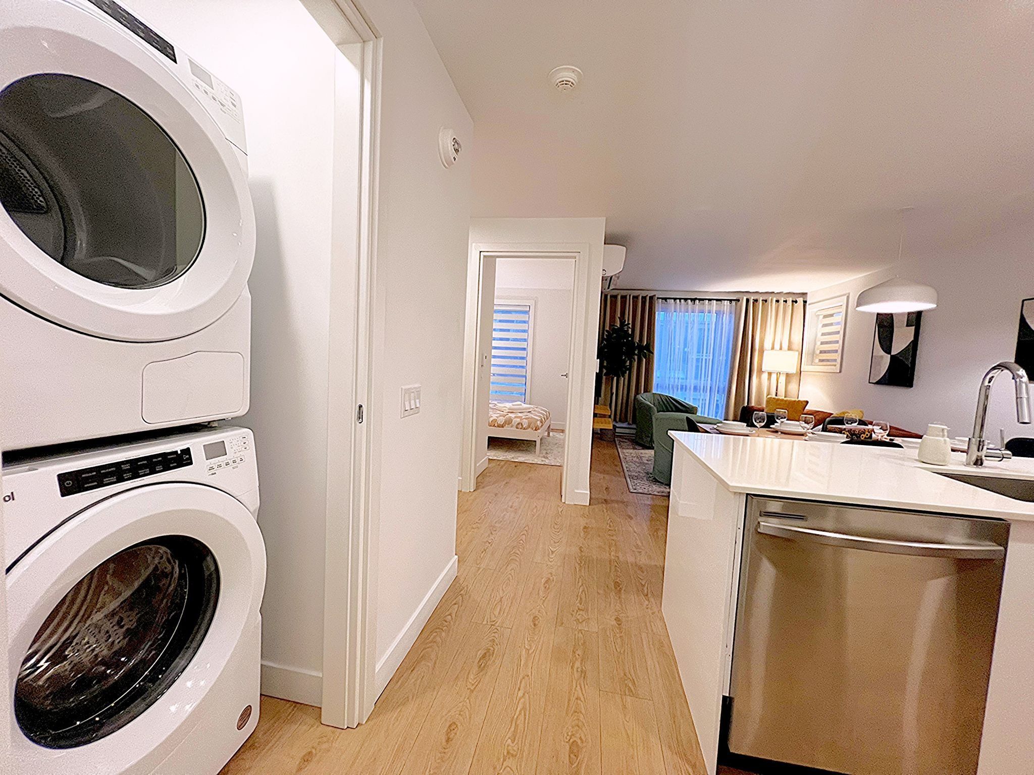 A compact hotel room with a washing machine, a small kitchenette, a dining area, and a bedroom with a window.