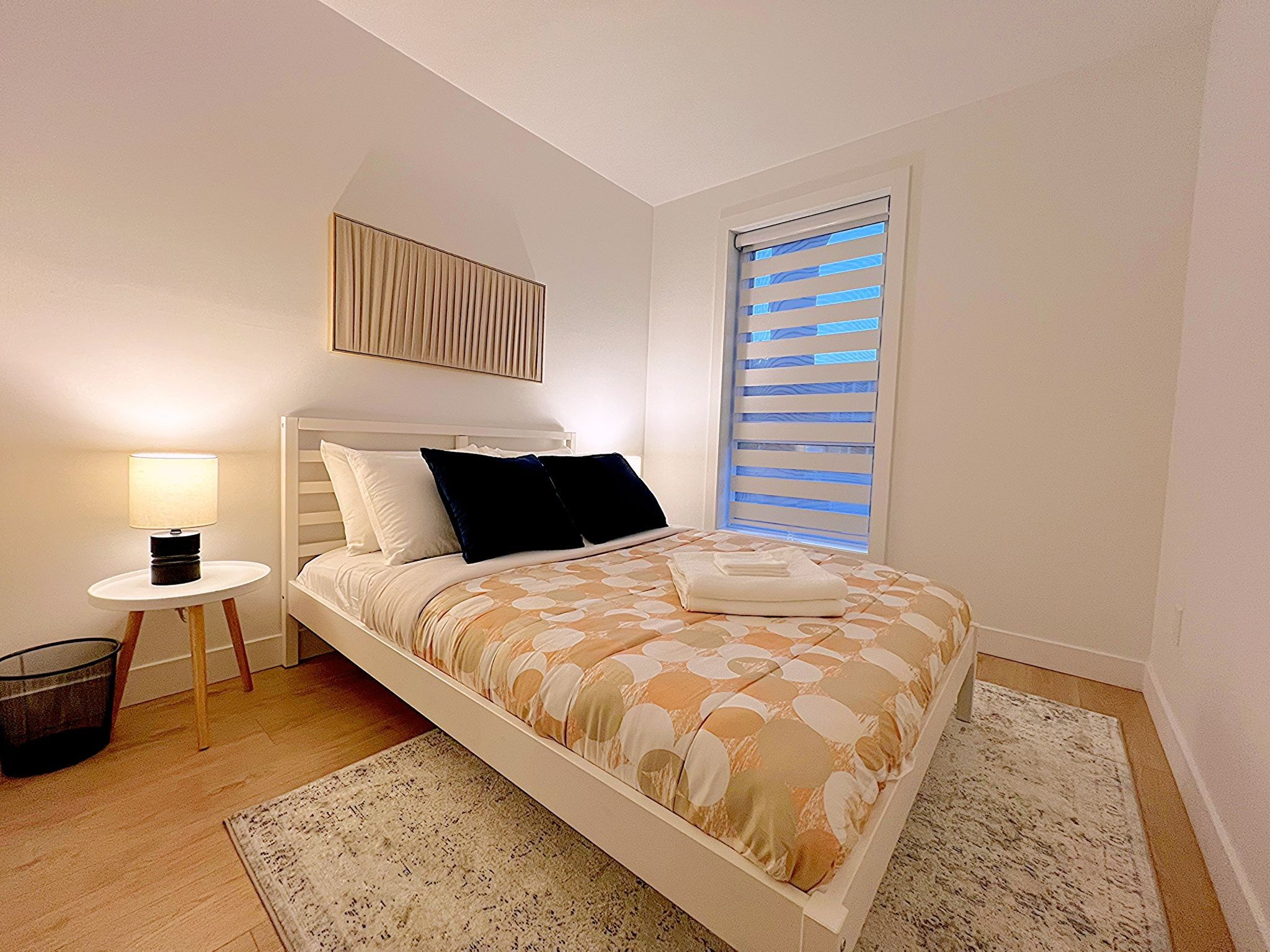 A bedroom with a bed, a bedside table with a lamp, a window with blinds, and a wall-mounted artwork.