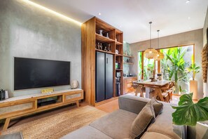 A modern living room with a flat-screen TV, wooden shelving, a dining area with a table and chairs, and a large window with plants. - Villa Dinova (Badung)