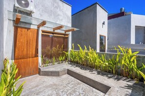 A modern building with a wooden door, a concrete pathway, and a row of green plants. - Villa Dinova (Badung)