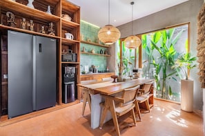 A modern kitchen with a built-in refrigerator, wooden cabinets, and a dining area with a large window overlooking greenery. - Villa Dinova (Badung)