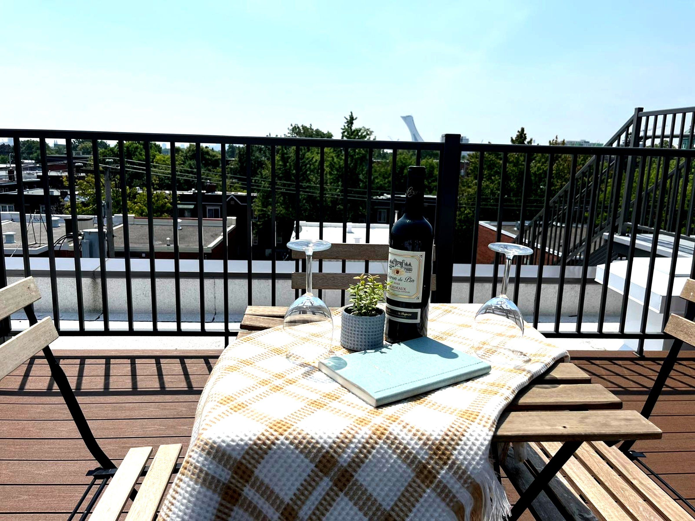 A table with a checkered tablecloth, a wine bottle, a glass, and a book on a balcony with a view of a parking area and trees.