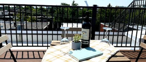 A table with a checkered tablecloth, a wine bottle, a glass, and a book on a balcony with a view of a parking area and trees.