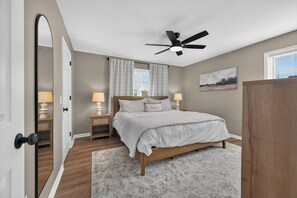 Une chambre à coucher avec un lit, deux tables de chevet, un ventilateur de plafond, une fenêtre avec des rideaux et un tableau au mur.