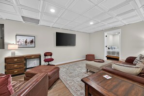 A spacious living room with a large flat-screen TV mounted on the wall, a wooden desk with drawers, a leather sofa, a red chair, and a patterned rug. - Four-bedroom Blacksburg home near Virginia Tech with mountain views, front and wraparound porches, fire pit, full kitchen, and basement air hockey. Features 2 king beds, twin room, basement bed, and loft twin beds across 3 levels. (Blacksburg)