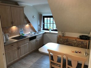 A compact kitchen with a dining area, a window with curtains, and a radiator. - Hansen Nieblum - Holiday Apartment 2 (Nieblum)