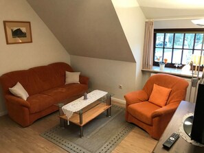 A living room with a brown sofa, a wooden coffee table, and an orange armchair. There is a window with curtains, a picture on the wall, and a remote control on the table. - Hansen Nieblum - Holiday Apartment 2 (Nieblum)