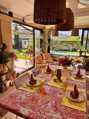 An indoor dining area with a table set for a meal, a view of the outdoors, and wicker furniture. - Une Bastide en Provence (Plan-d'Orgon)