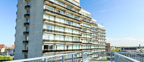 A multi-story residential building with balconies, a pedestrian bridge, and a clear sky.