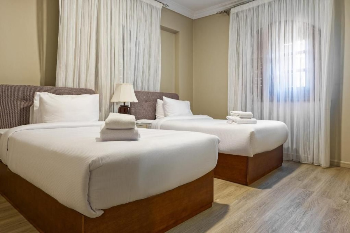 A hotel room with two beds, a nightstand, a lamp, and a window with curtains.
