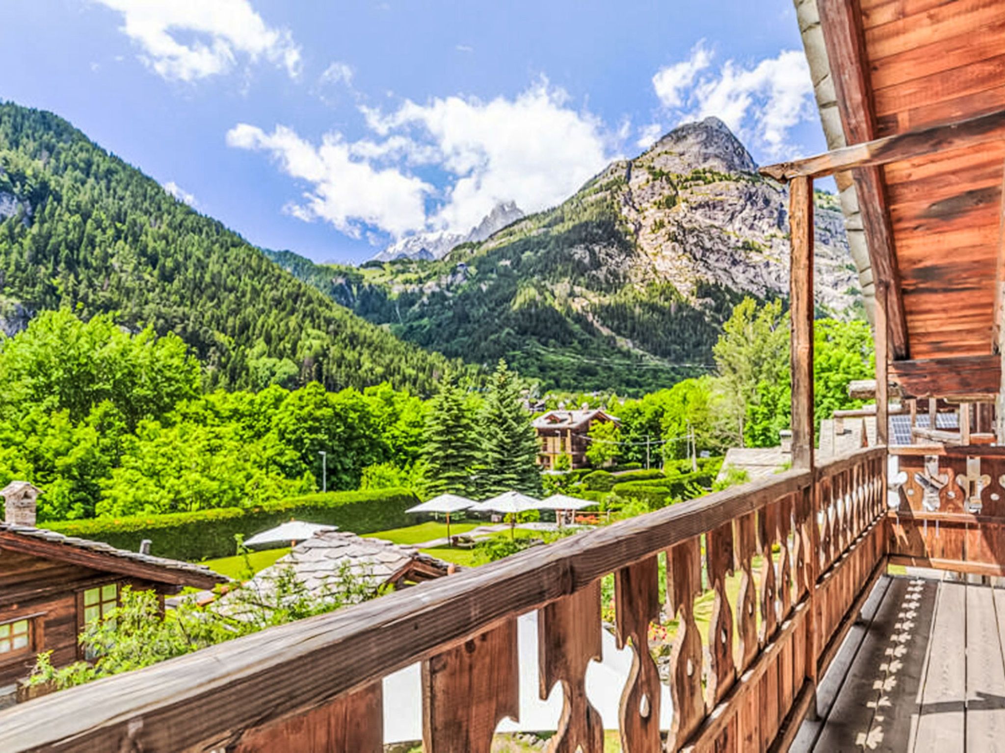A wooden balcony with a view of a mountainous landscape, a lake, and a building with a sloped roof.