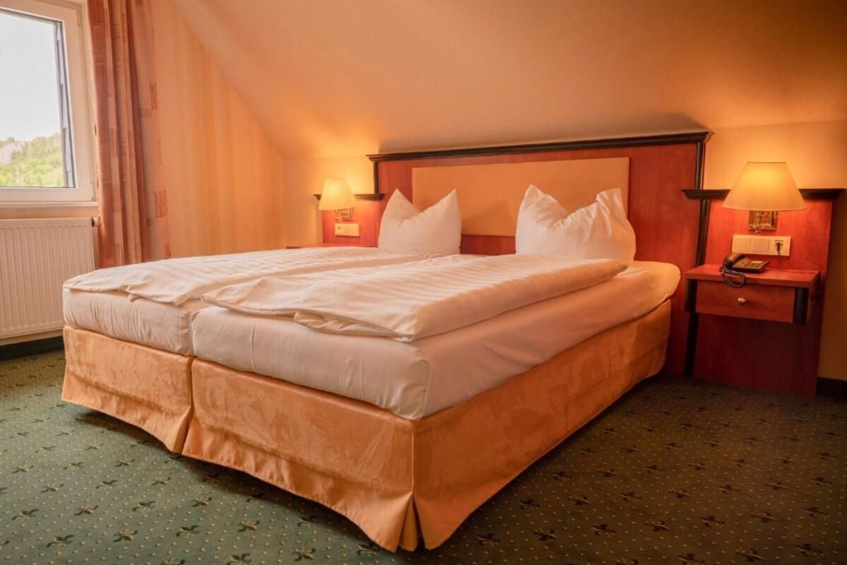 A hotel room with a large bed, two bedside tables with lamps, a window with curtains, and a carpeted floor.