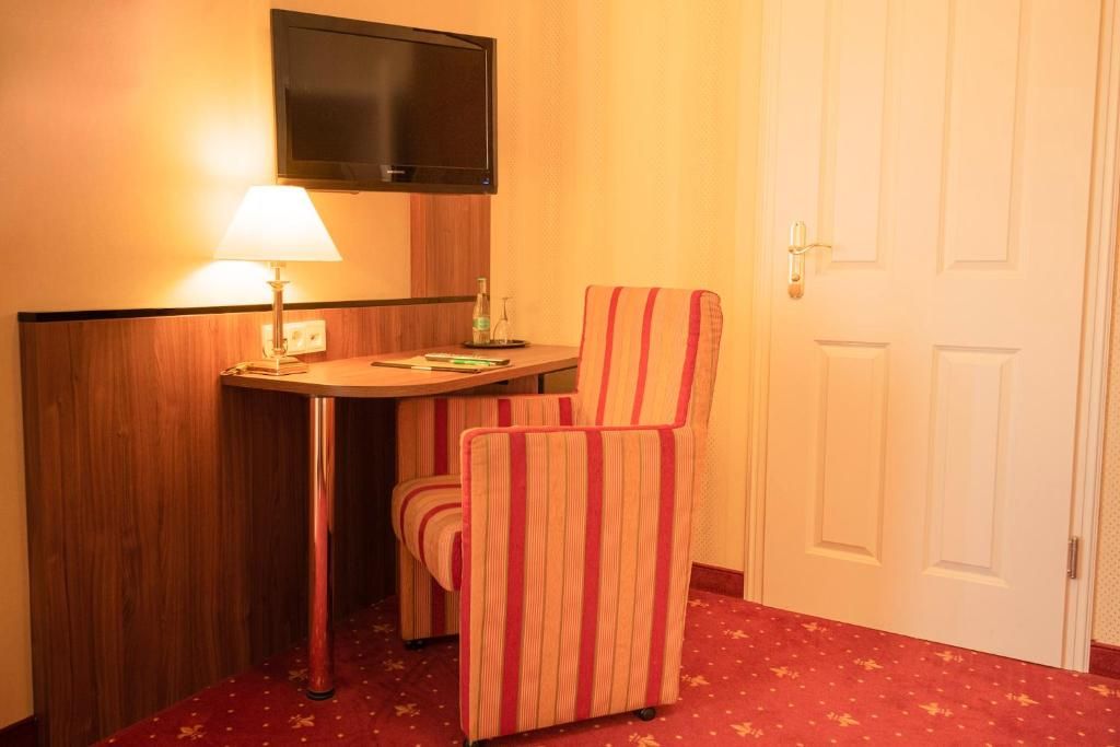 Una habitación de hotel con una cama individual, una mesita de noche con lámpara, un televisor montado en la pared y un sillón a rayas.
