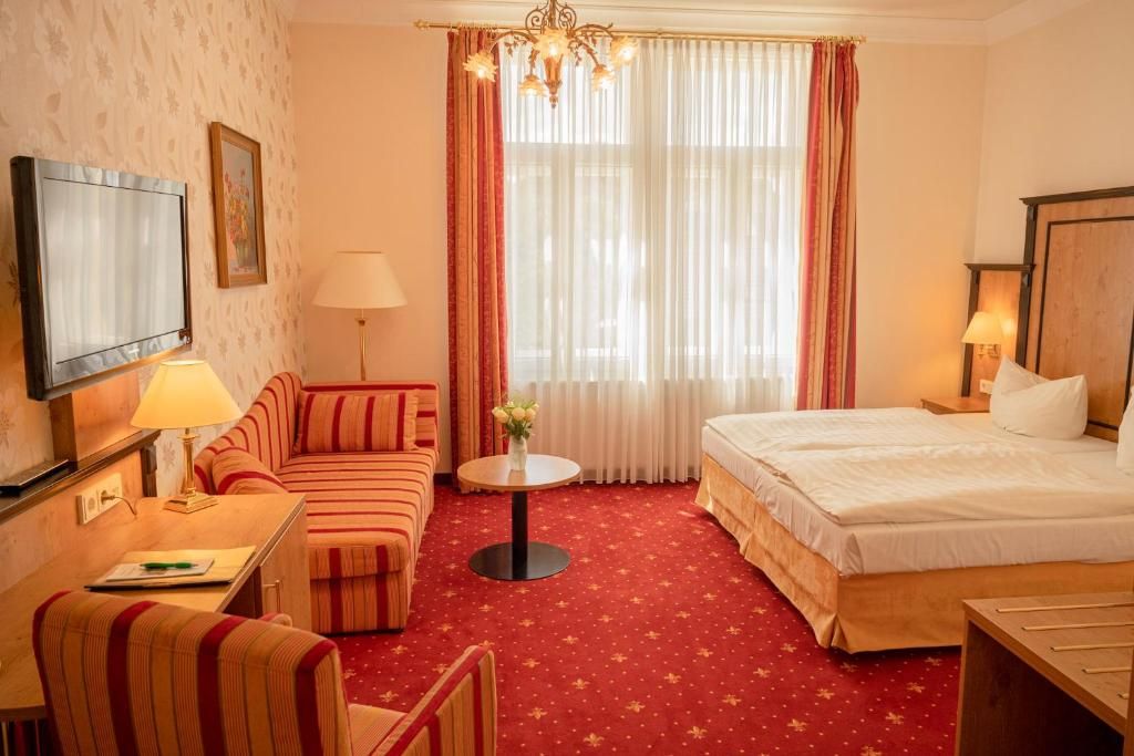 A hotel room with a bed, a sofa, a desk with a lamp, a television, and a window with curtains.