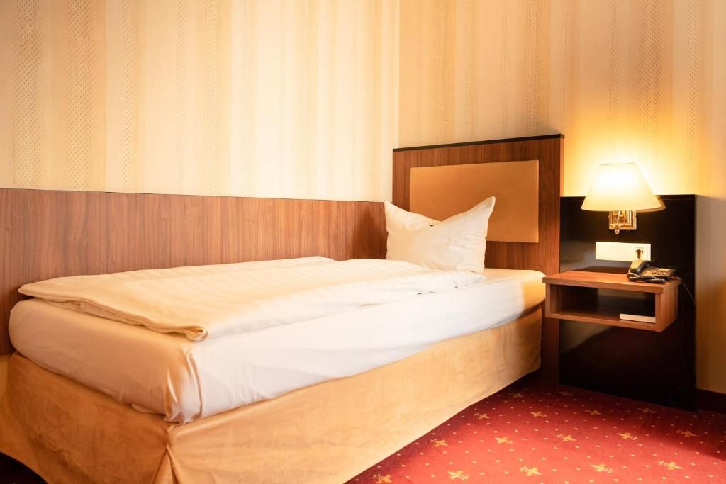 A hotel room with a bed, a nightstand with a lamp, and a red carpet.