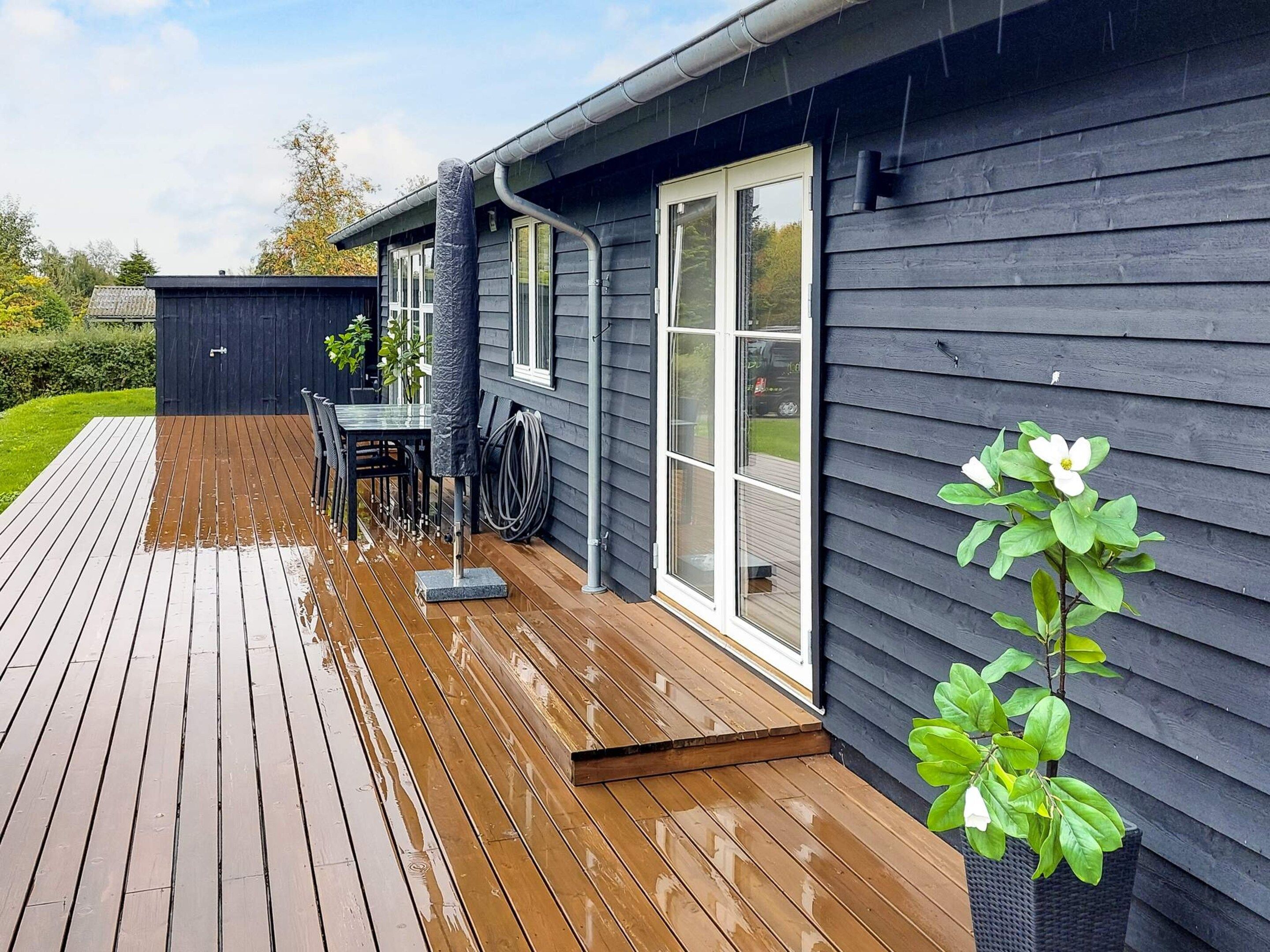 A wooden deck with a table and chairs, a covered patio area, and a house with dark siding and a large window.