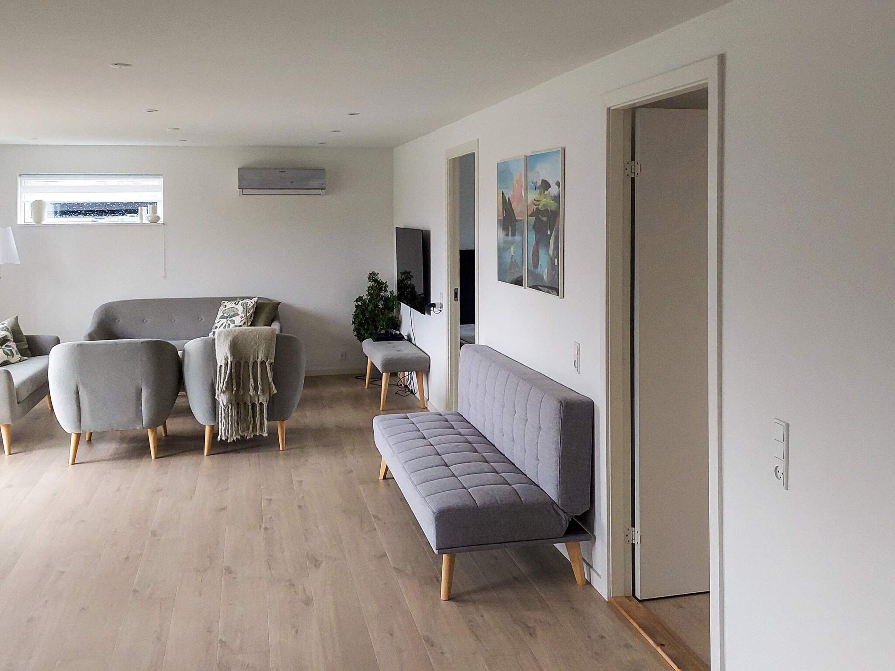 A modern living room with a grey sofa, two armchairs, a bench, and a flat-screen TV mounted on the wall.
