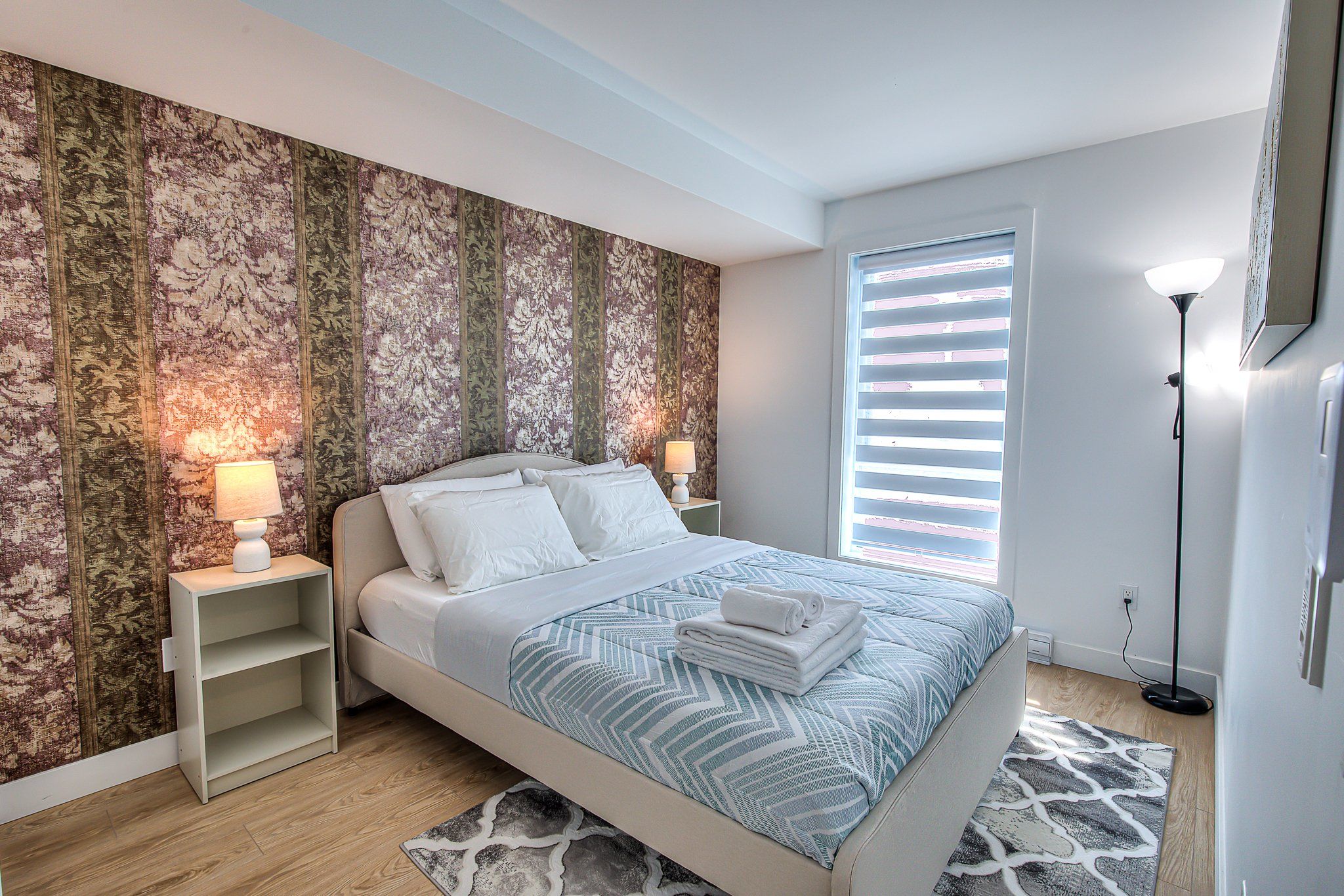 A bedroom with a bed, nightstands, a lamp, a window with blinds, and a patterned rug.