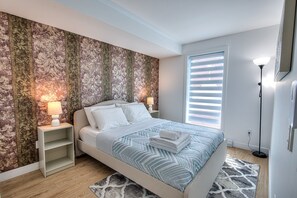 A bedroom with a bed, nightstands, a lamp, a window with blinds, and a patterned rug. - Elegant 3BR Condo for Families | 2 Baths, AC & Fast WiFi (Montréal)