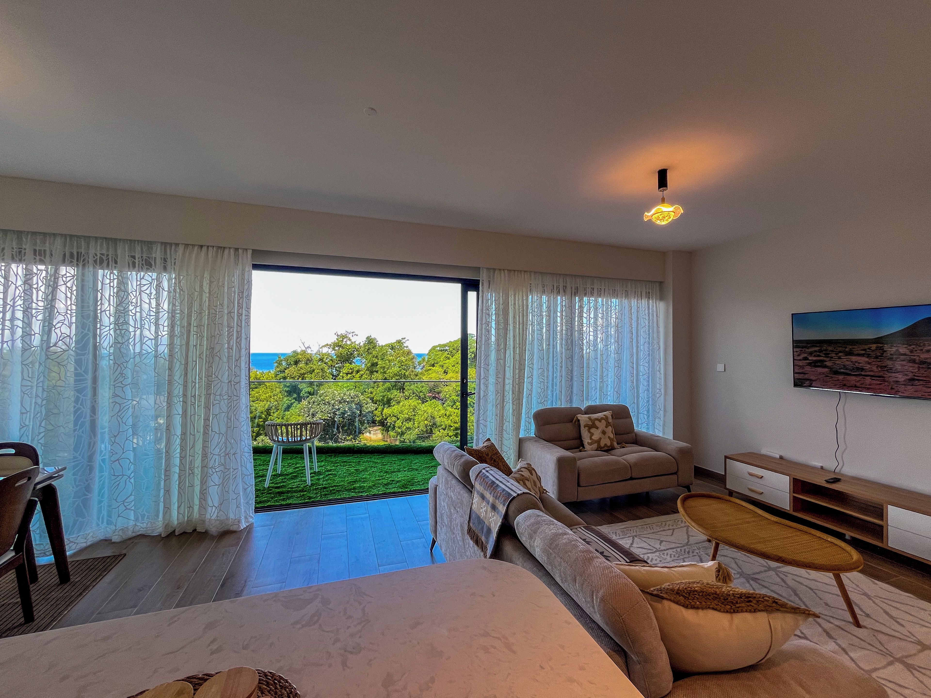 A modern bedroom with a large bed, a sofa, a coffee table, and a TV. There is a balcony with a view of the sea and trees.