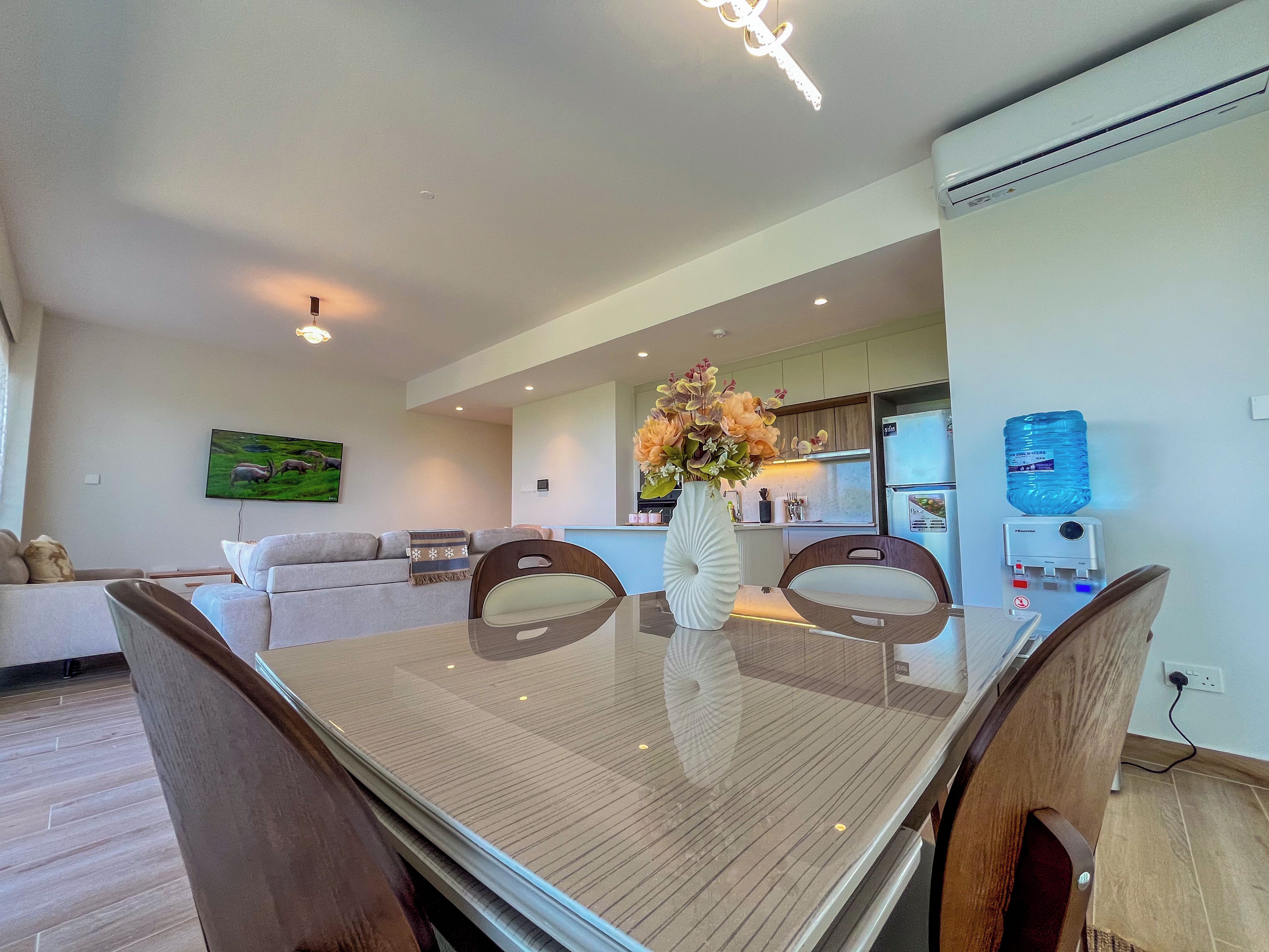 A modern living room with a glass dining table, a sofa, and a kitchen area in the background.