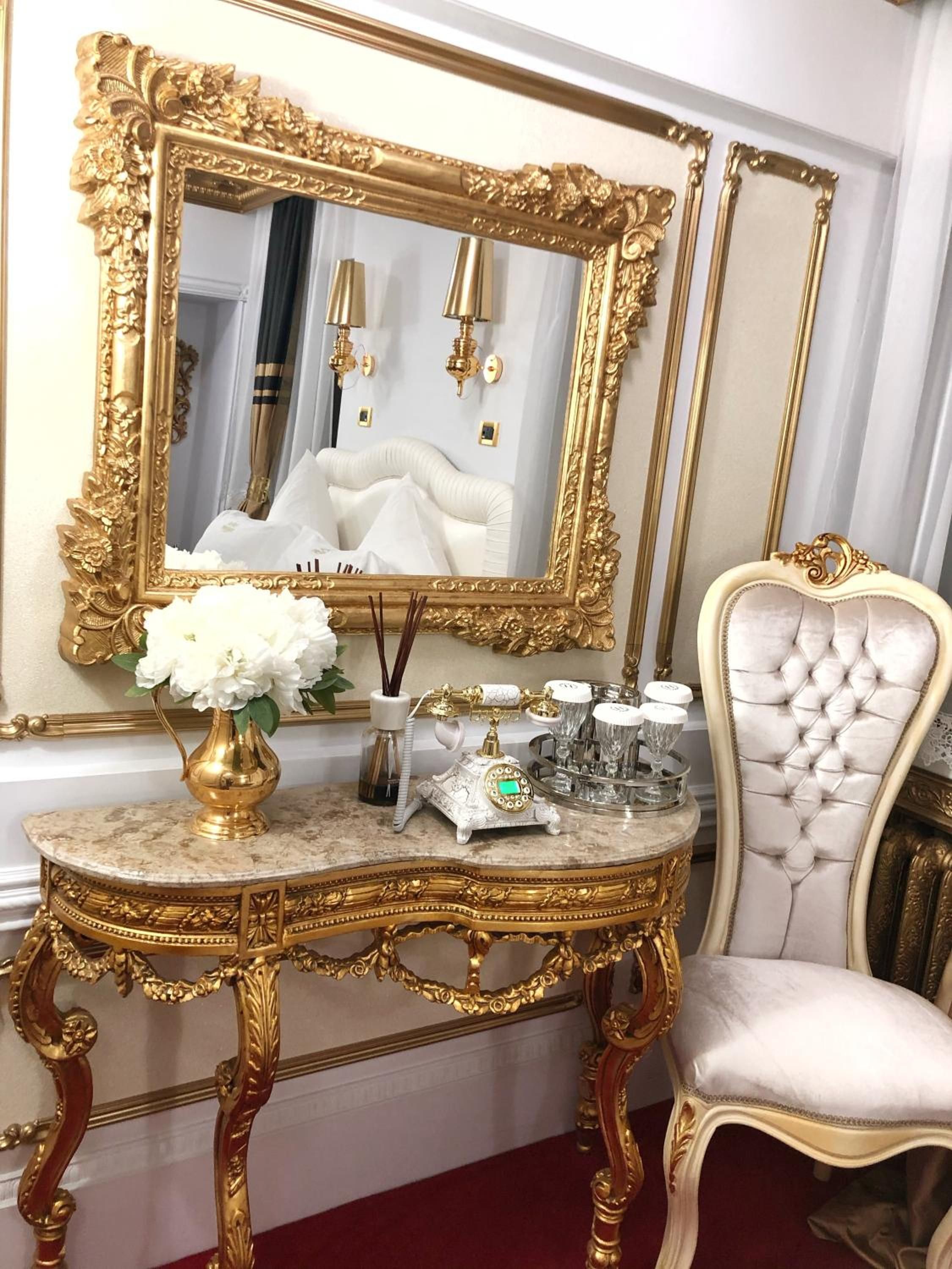 Quarto luxuoso com toucador de mármore, espelho com moldura dourada, poltrona estofada em branco e um vaso com flores brancas.