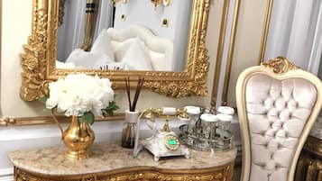 Quarto luxuoso com toucador de mármore, espelho com moldura dourada, poltrona estofada em branco e um vaso com flores brancas.