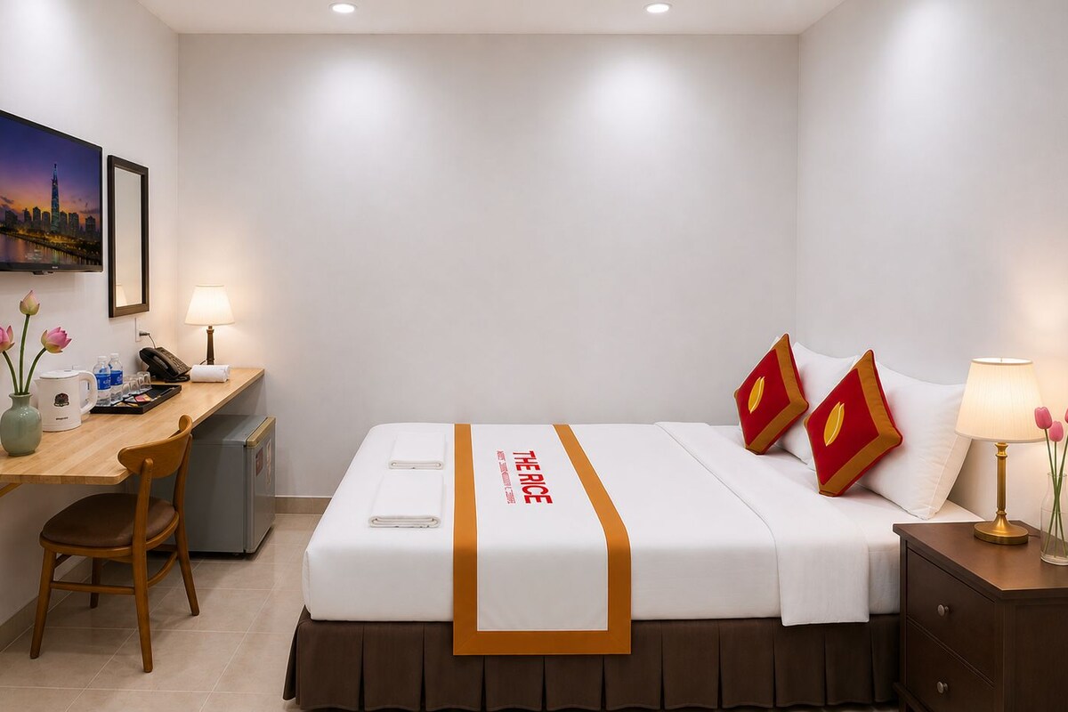 A hotel room with a bed, desk, chair, and a nightstand with a lamp.