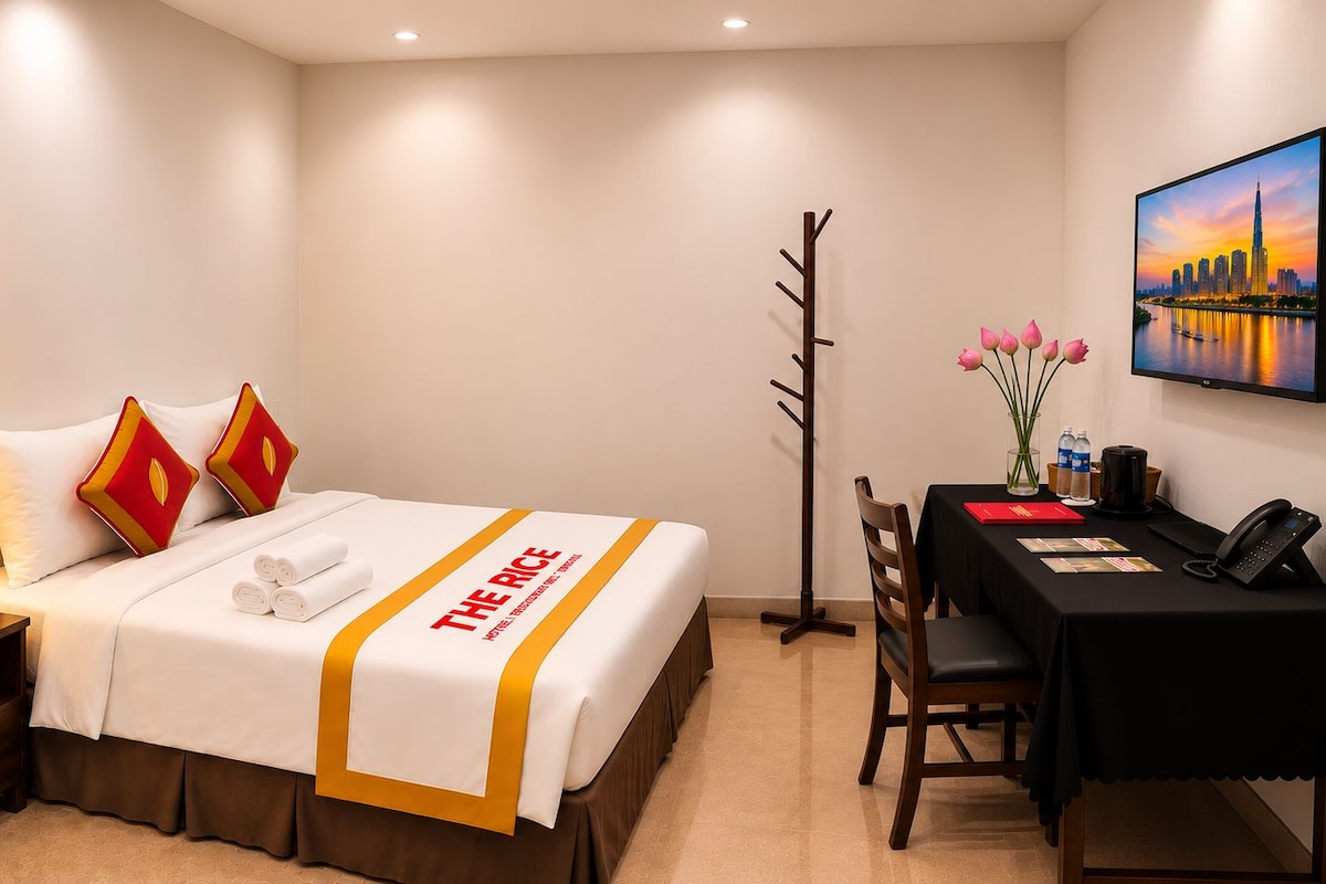 A hotel room with a bed, a desk, a chair, a lamp, a coat rack, and a painting of a cityscape.