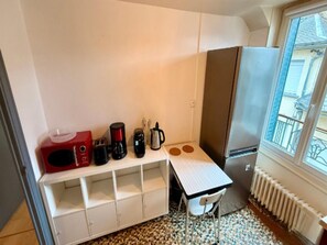 Eine kompakte Küchenzeile mit Mikrowelle, Kaffeemaschine, Toaster und Wasserkocher auf einem weißen Regal, ein kleiner Tisch mit Stuhl und ein Kühlschrank.