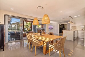 A modern kitchen with a central island, a dining area with a wooden table and wicker chairs, and a large sliding glass door leading to an outdoor area. - Burleigh Waters Luxury Getaway - Pool & Lake Views (Burleigh Waters)