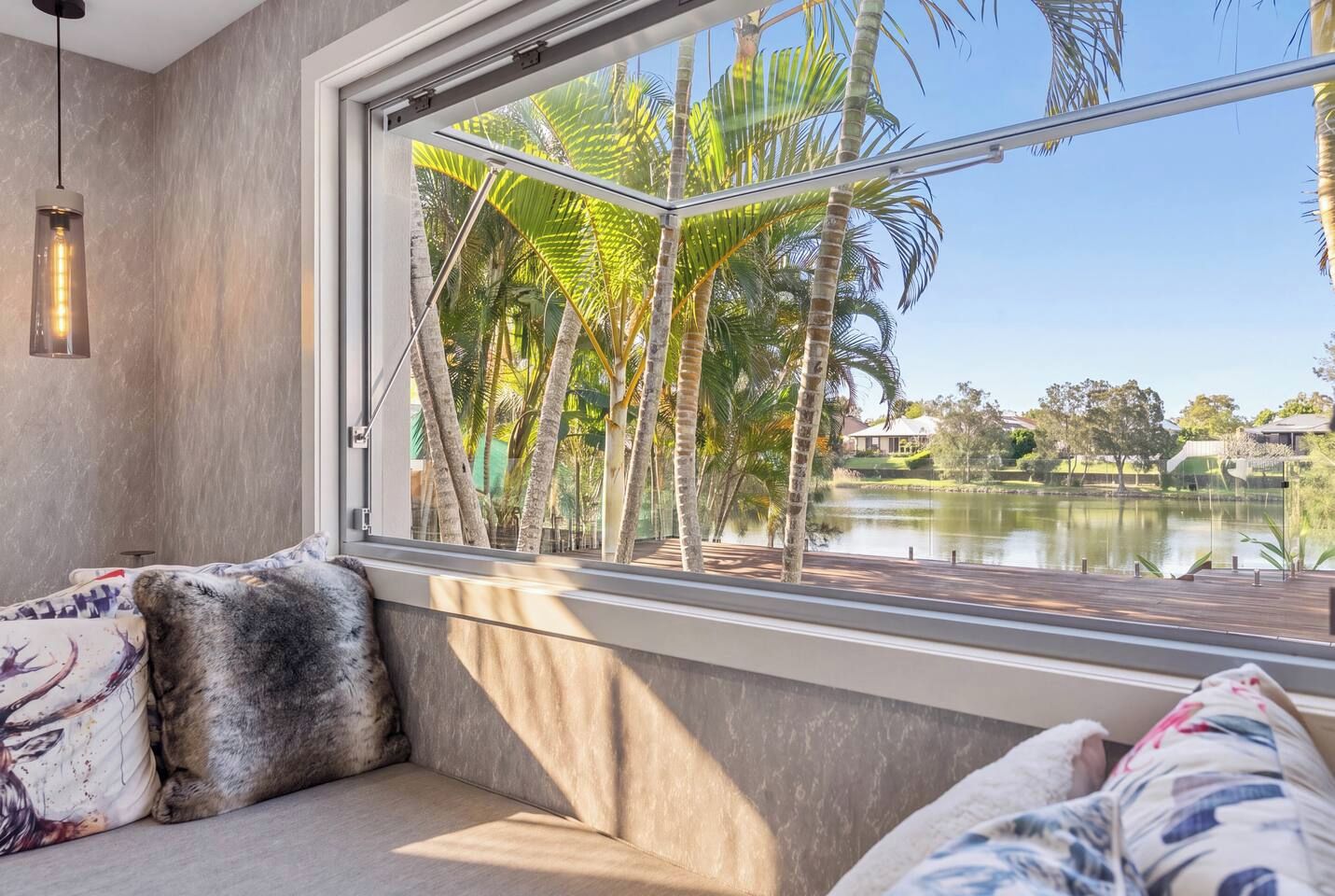 Una habitación con un ventanal que da a un lago y palmeras, un sofá con cojines estampados y una lámpara colgante.