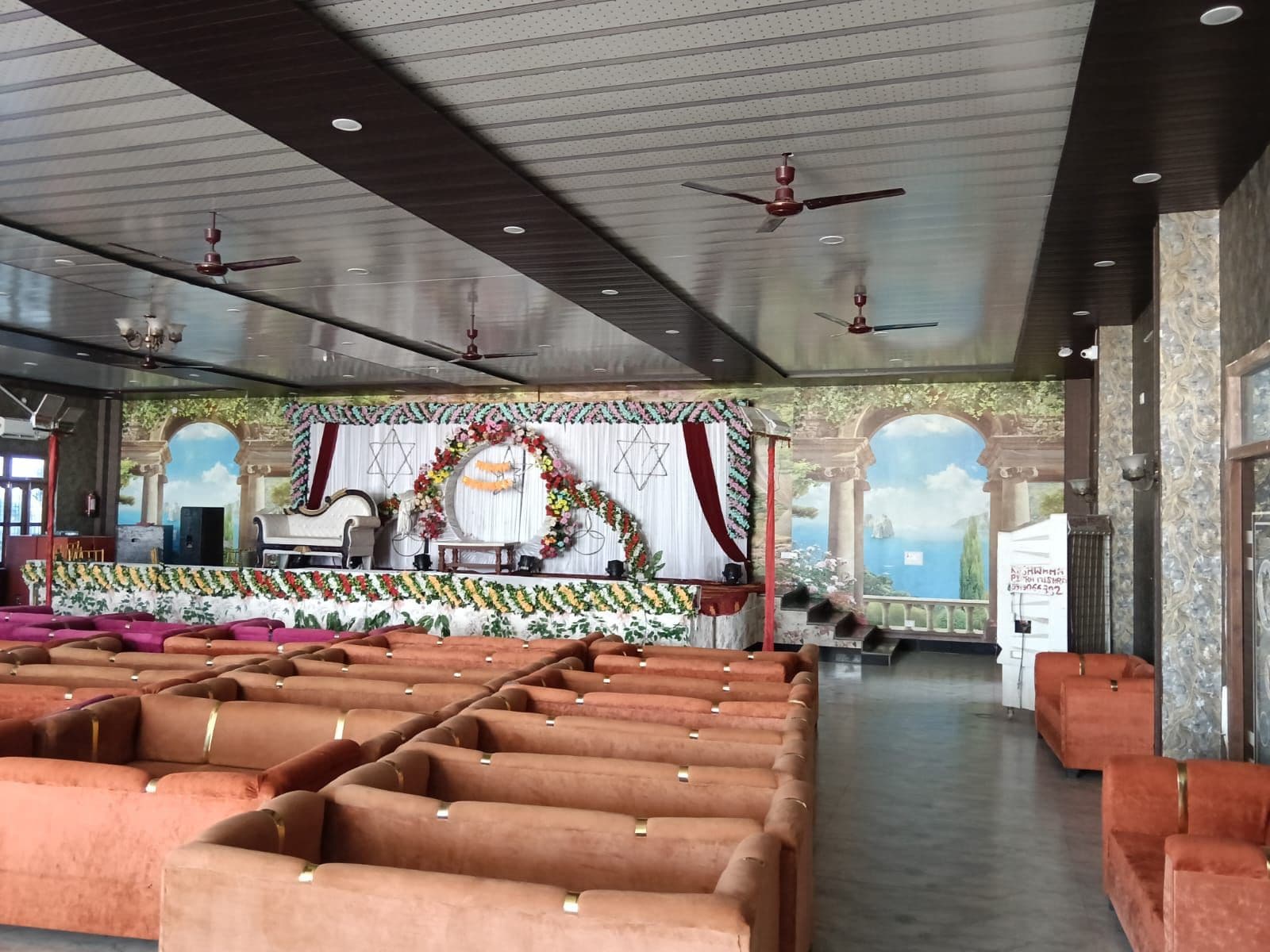 A spacious hall with rows of orange sofas, a stage with floral decorations, and ceiling fans.