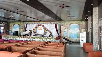 A spacious hall with rows of orange sofas, a stage with floral decorations, and ceiling fans.