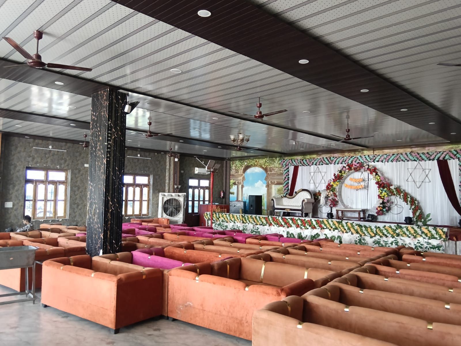 A spacious hall with rows of orange sofas, a stage with floral decorations, and a ceiling with recessed lighting.