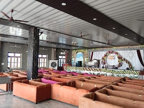 A spacious hall with rows of orange sofas, a stage with floral decorations, and a ceiling with recessed lighting. - Wishotel Resort Rajkumari Hridya Palace  (Padri Gajarat)