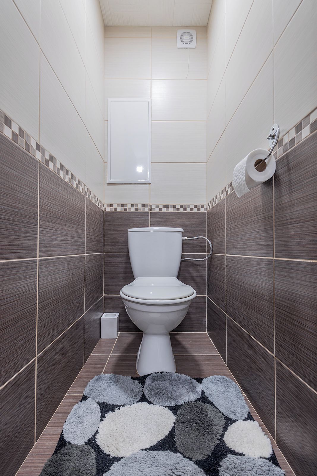 A bathroom with a toilet, a small trash bin, a towel rack, and a patterned rug on the floor.