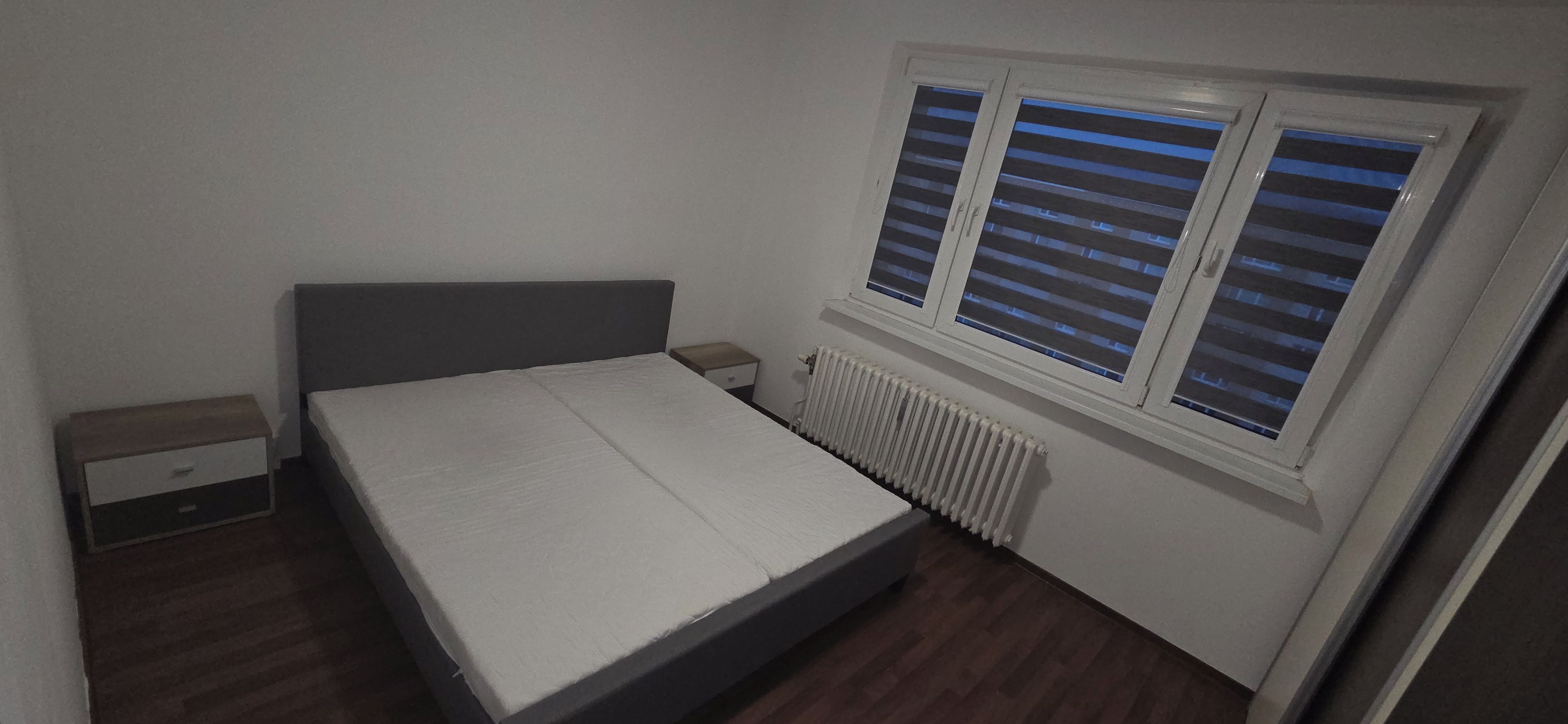 A small, single-bed room with a wooden floor, a white mattress, a nightstand, a drawer, and a window with blinds.