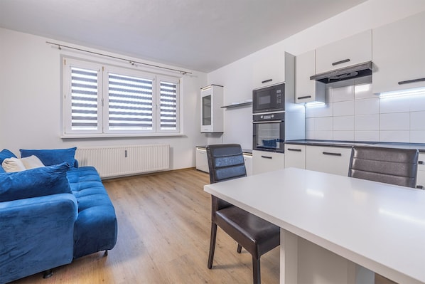 A modern kitchen with white cabinets, a built-in oven, and a microwave. There is a dining table with chairs, a blue sofa with cushions, and a window with blinds.