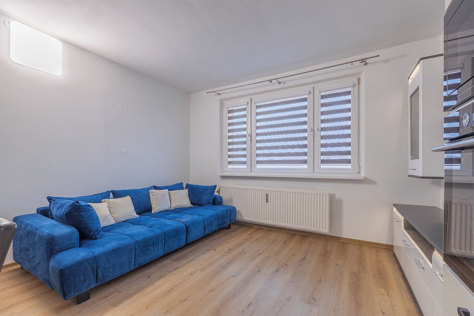 A room with a blue sofa, a radiator, a window with blinds, and a kitchenette area.