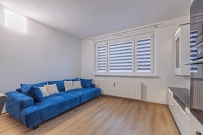 Ein Zimmer mit einem blauen Sofa, einem Heizkörper, einem Fenster mit Jalousien und einer Küchenzeile.