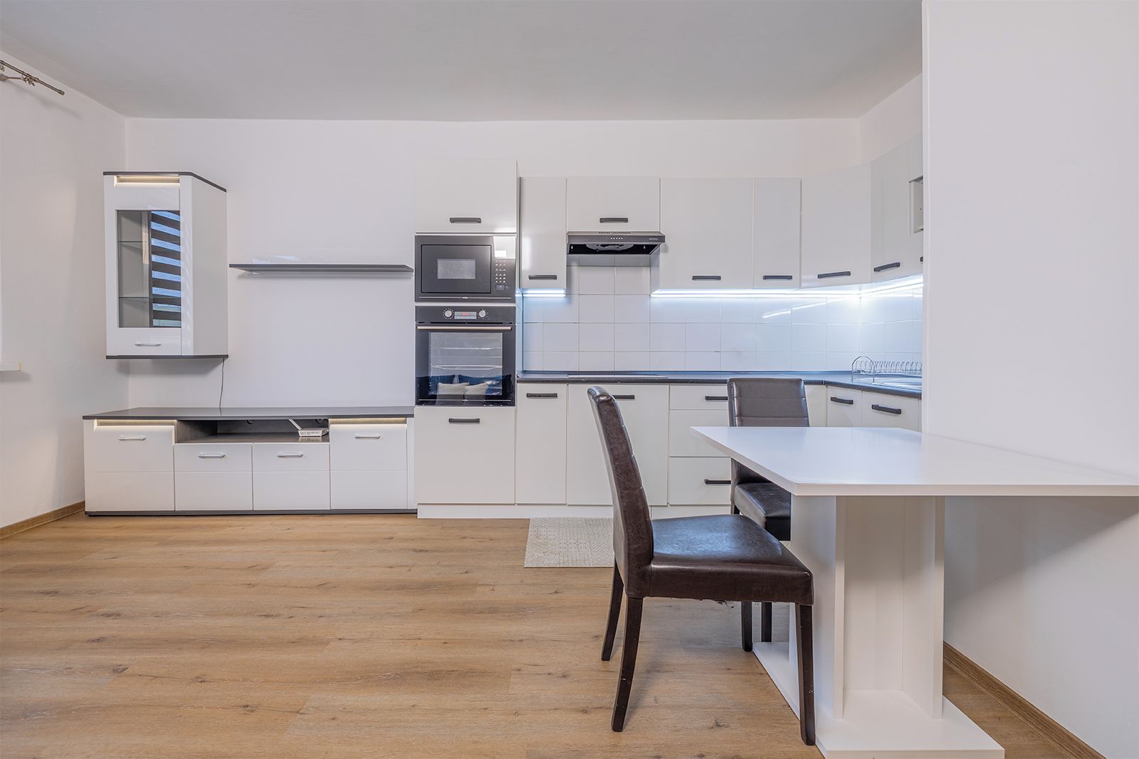 Una cocina moderna con armarios blancos, horno empotrado y comedor con mesa y sillas.