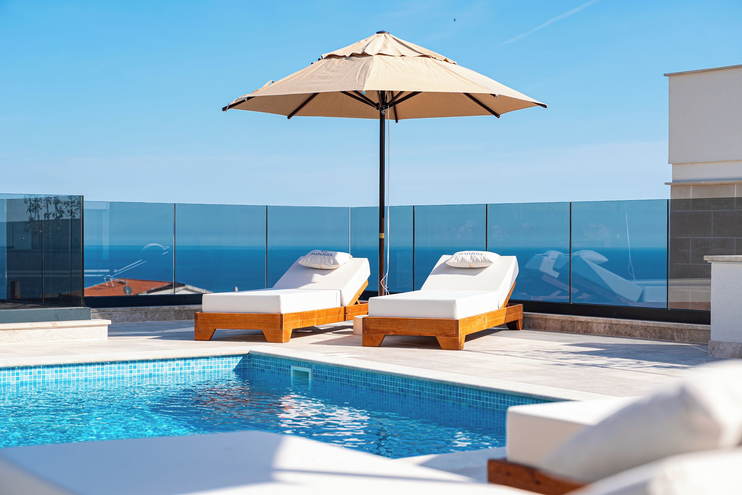 A rooftop pool area with two lounge chairs, a large umbrella, and a clear view of the ocean.