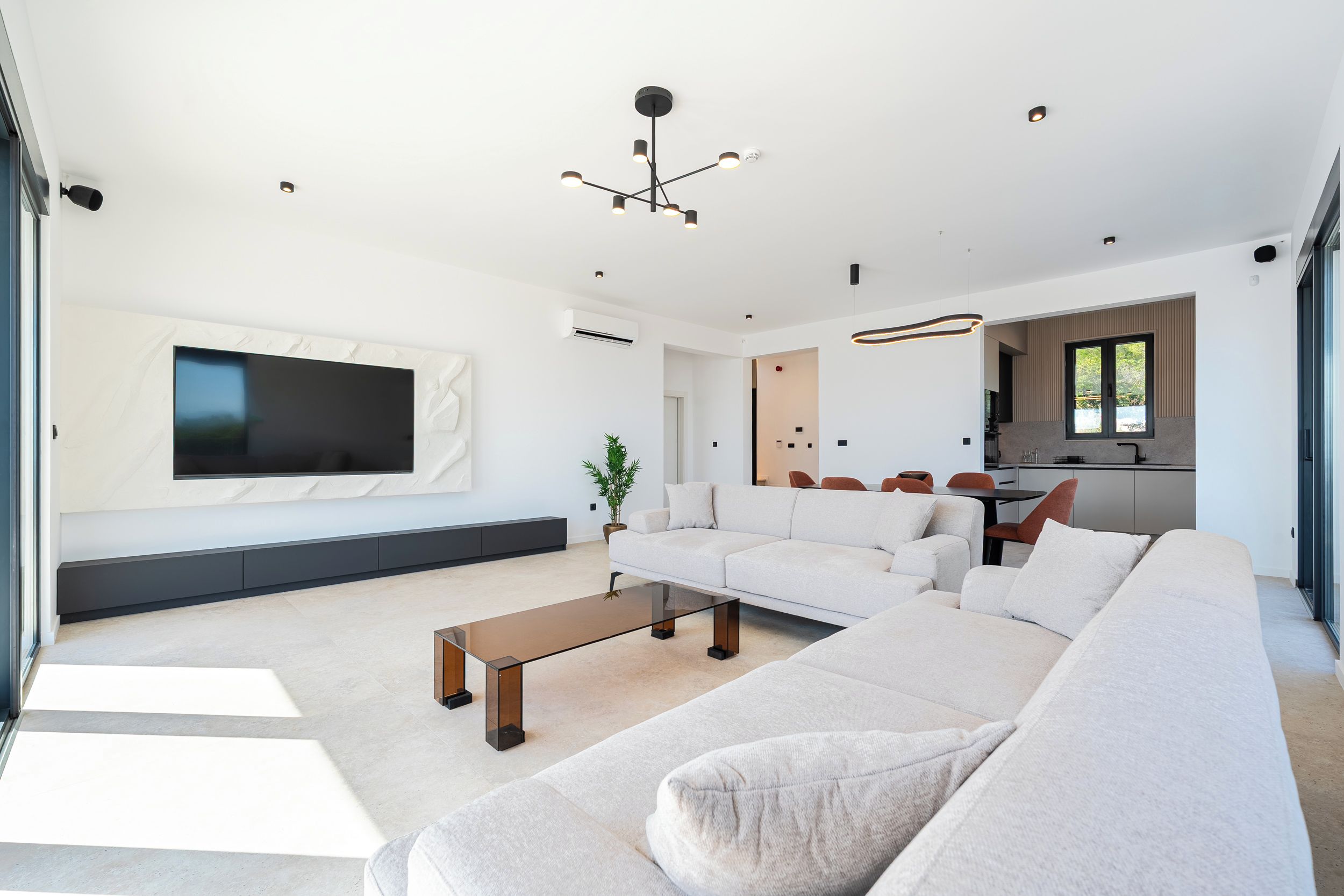 A modern living room with a large sectional sofa, a flat-screen TV mounted on the wall, a coffee table, and a kitchen area visible through an open door.