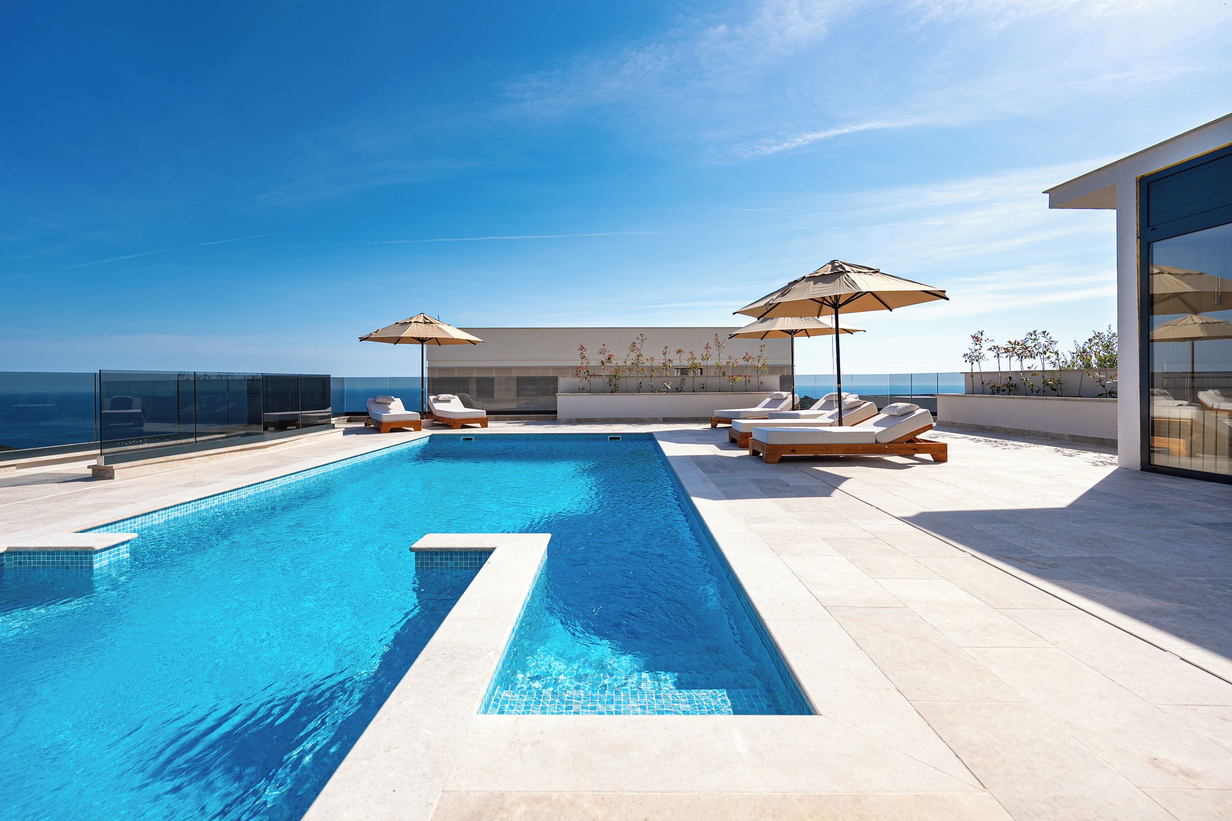 En modern infinitypool med kristallklart blått vatten, omgiven av solstolar och parasoller, med utsikt över en klar himmel och en avlägsen horisont.