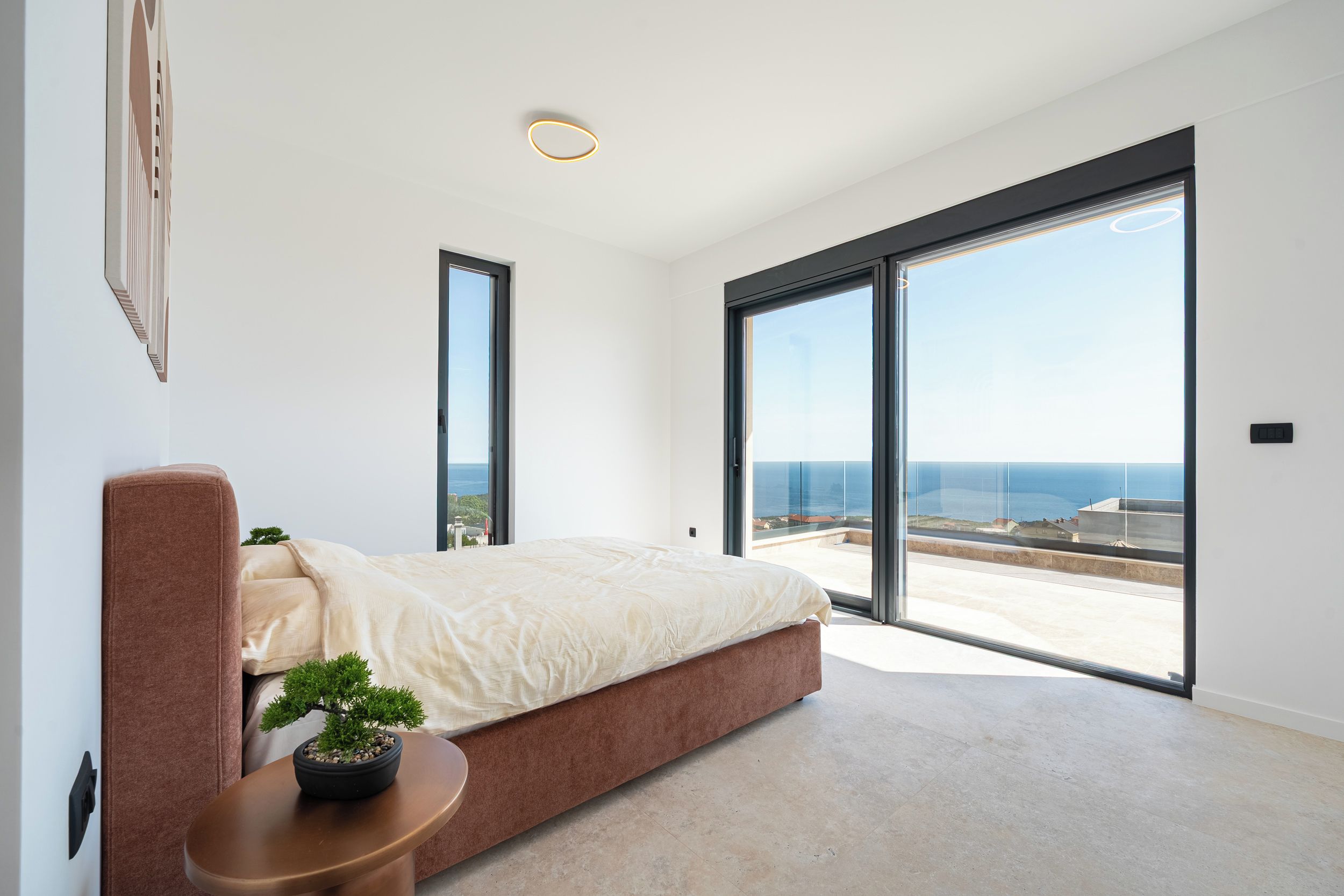A modern bedroom with a large bed, a small side table, a potted plant, and a view of the sea through large sliding doors.