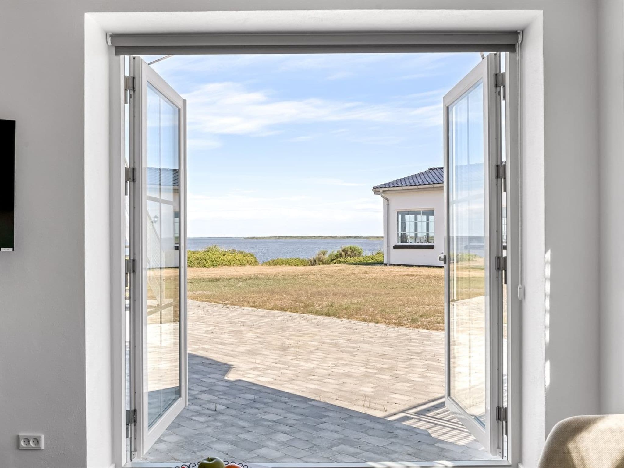 A view through a glass door leading to a paved outdoor area with a building in the background and a body of water.