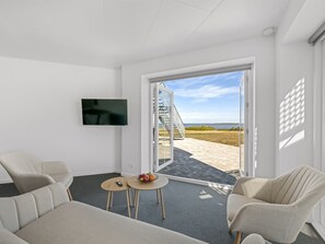 A modern hotel room with a flat-screen TV, a sofa, two chairs, a small table with fruit, and a view of the beach through a glass door. - "Alban" - 50m to the fjord by Interhome (Tarm)